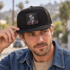 A man wearing a black trucker hat with a skull patch on the front, holding the brim. The background is a softly blurred outdoor street scene.
