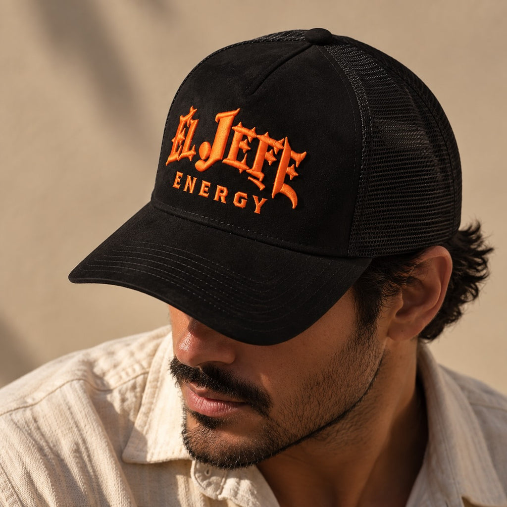 A man wearing a black trucker hat with orange embroidered text reading 'El Jefe Energy' on the front, photographed in warm natural light against a beige wall.