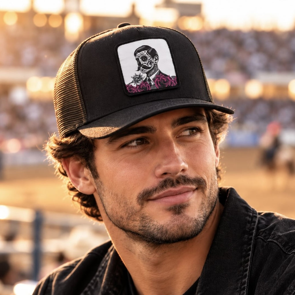 A man wearing a black trucker hat with a white square patch featuring a black and white skull illustration with pink roses, set against a blurred outdoor stadium background.