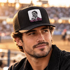 A man wearing a black trucker hat with a white square patch featuring a black and white skull illustration with pink roses, set against a blurred outdoor stadium background.