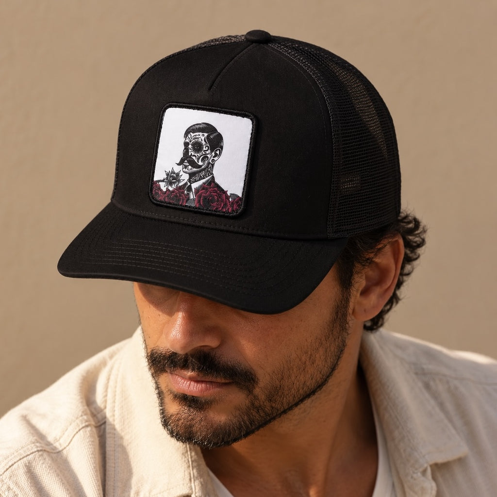 A man wearing a black trucker hat with a square embroidered patch on the front. The patch features a white background with a black-and-white portrait of El Jefe, with red roses along the bottom.