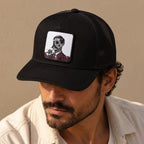 A man wearing a black trucker hat with a square embroidered patch on the front. The patch features a white background with a black-and-white portrait of El Jefe, with red roses along the bottom.