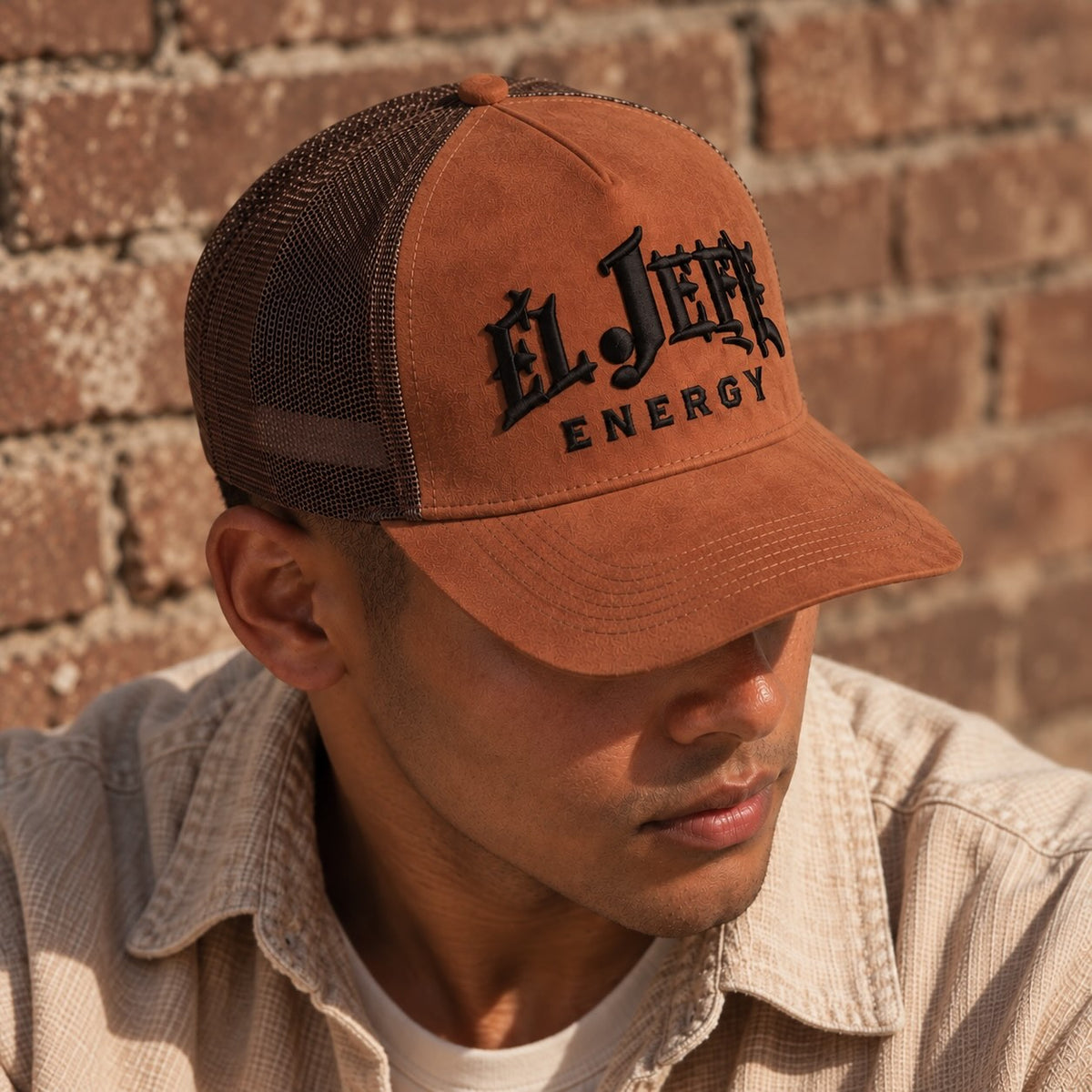 A man wearing a brown and black trucker hat with 'El Jefe Energy' embroidered on the front, set against a rustic brick wall background.