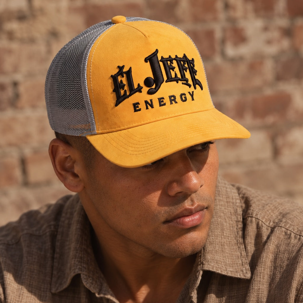 A man wearing a yellow and gray trucker hat with 'El Jefe Energy' embroidered on the front, set against a rustic brick wall background.