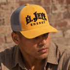 A man wearing a yellow and gray trucker hat with 'El Jefe Energy' embroidered on the front, set against a rustic brick wall background.