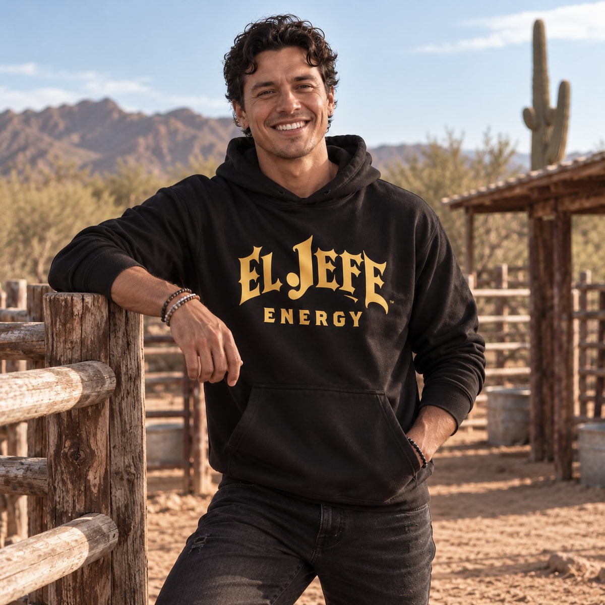 A man wearing a black pullover hoodie with 'EL JEFE ENERGY' printed in gold across the chest, posing outdoors in a desert ranch setting with mountains and a wooden fence in the background.