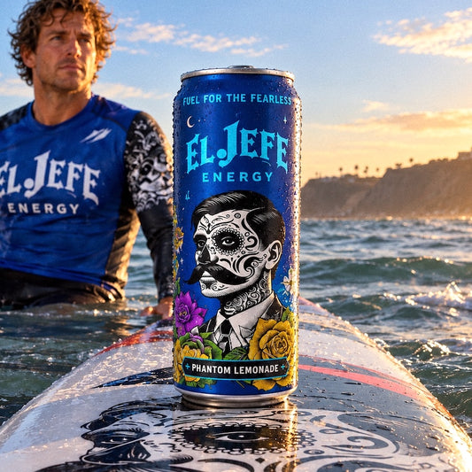 A blue El Jefe Energy can labeled Phantom Lemonade stands on a wet surfboard in the ocean, with a man in a blue rash guard standing in the water behind it during sunset.