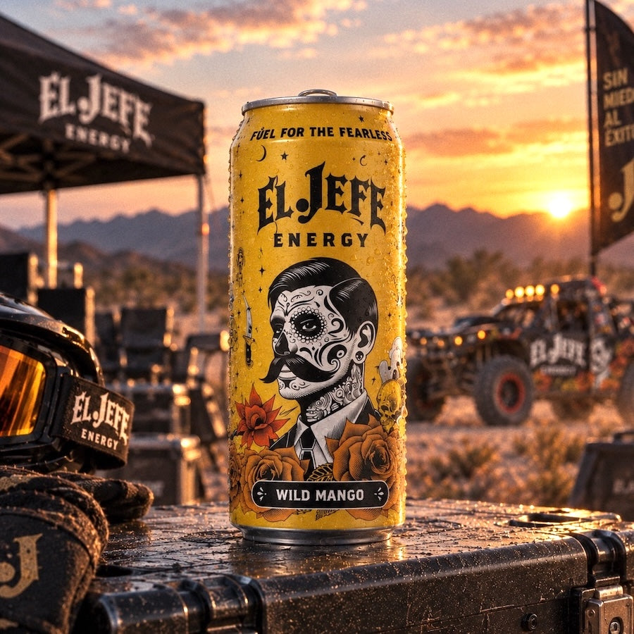 A yellow El Jefe Energy can labeled Wild Mango stands on a dark surface at sunset, with off-road vehicles, mountains, and tents in the background. The can features a black-and-white illustrated face with decorative text and floral artwork.
