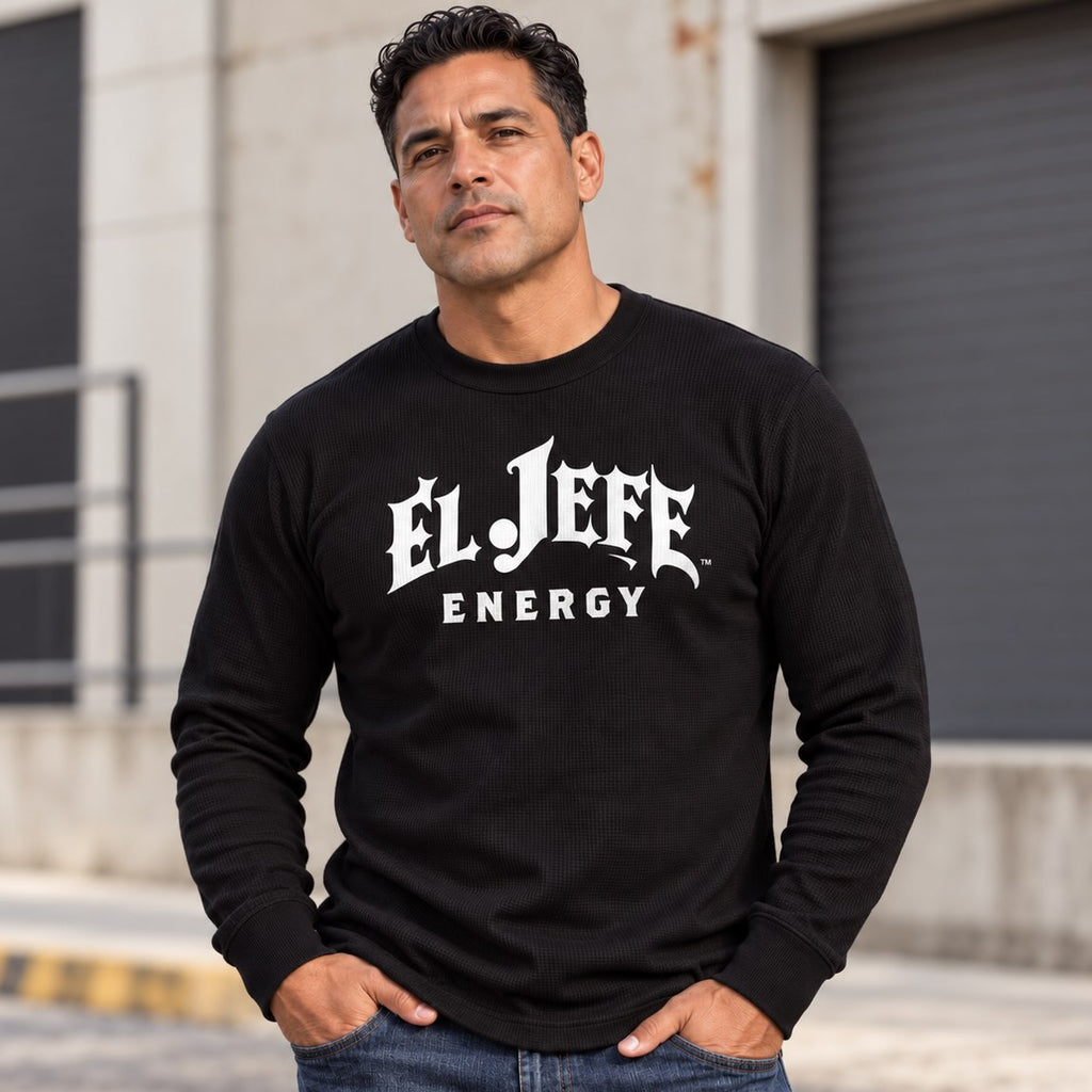 A man wearing a black long-sleeve waffle-knit t-shirt with 'EL JEFE ENERGY' printed in white gothic and sans-serif fonts on the chest, standing outdoors in front of a concrete building.