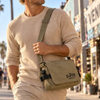 A man wearing a beige crewneck sweater and khaki pants carries a beige canvas messenger bag with black straps and the brand name 'EL JEFE ENERGY' printed on the front, set against a blurred city street background.