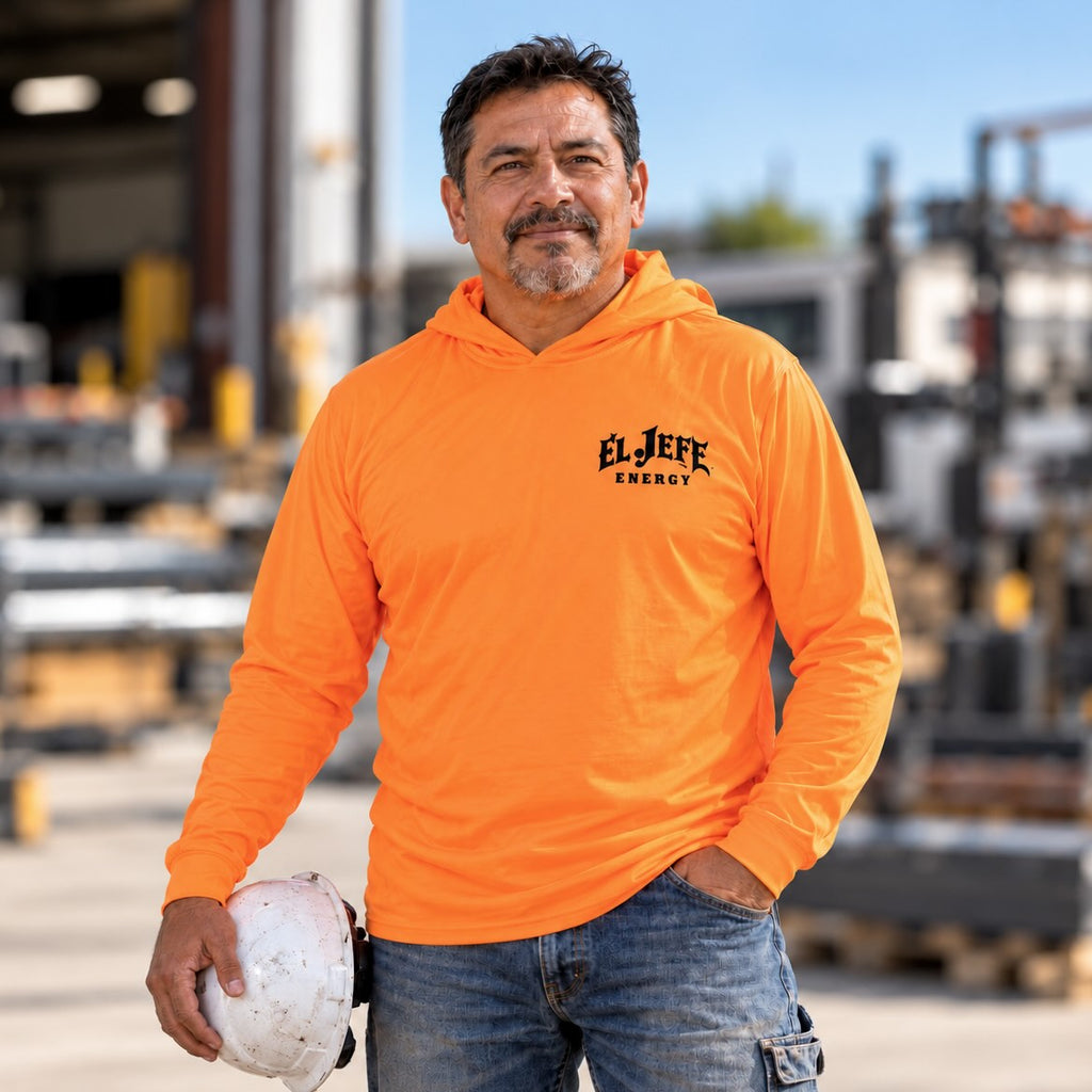 A man wearing a bright orange long-sleeve hoodie with 'EL JEFE ENERGY' printed in black on the left chest, standing in an industrial outdoor setting and holding a white hard hat.
