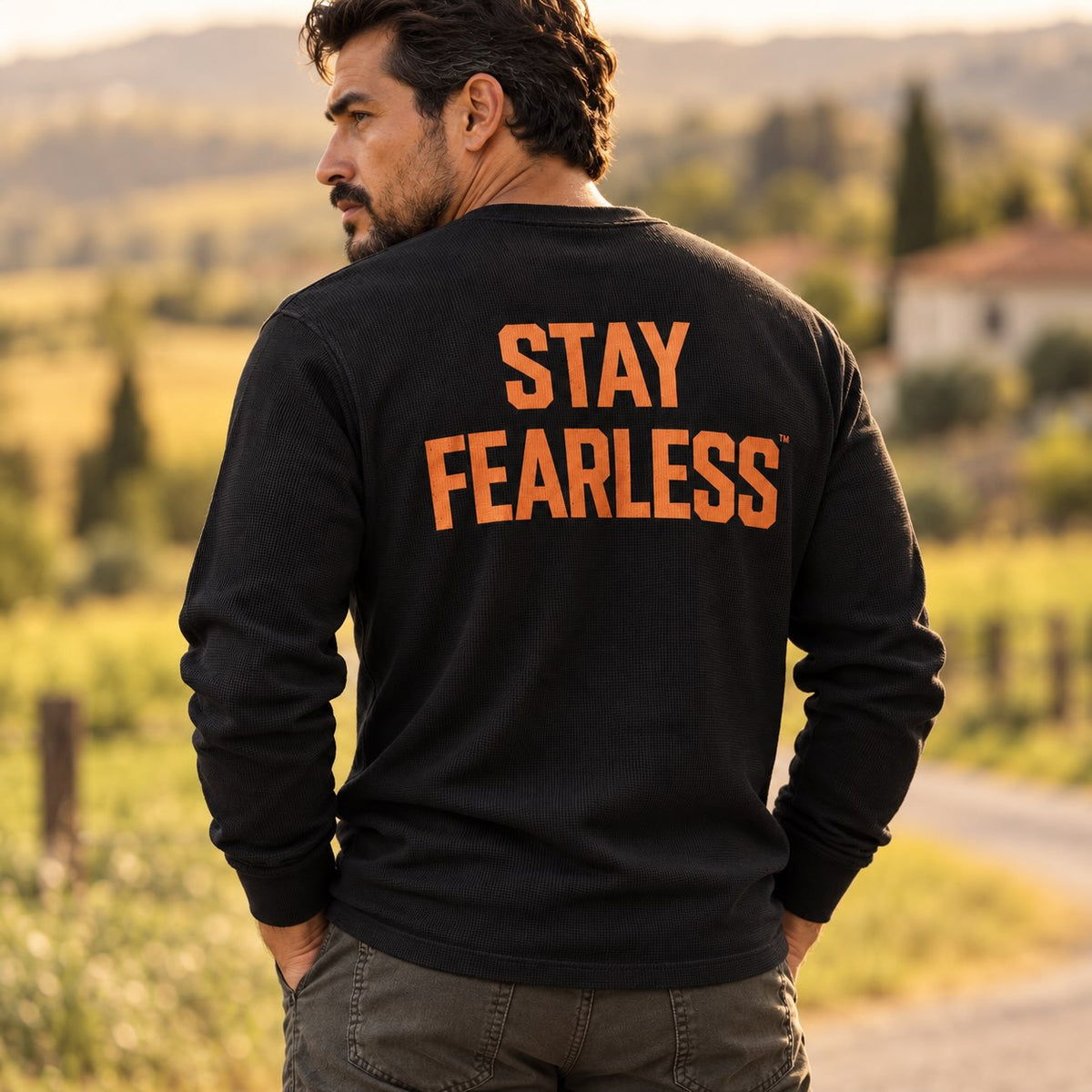 A man wearing a black long-sleeve sweatshirt with 'STAY FEARLESS' printed in orange across the back, photographed outdoors in a blurred rural landscape.