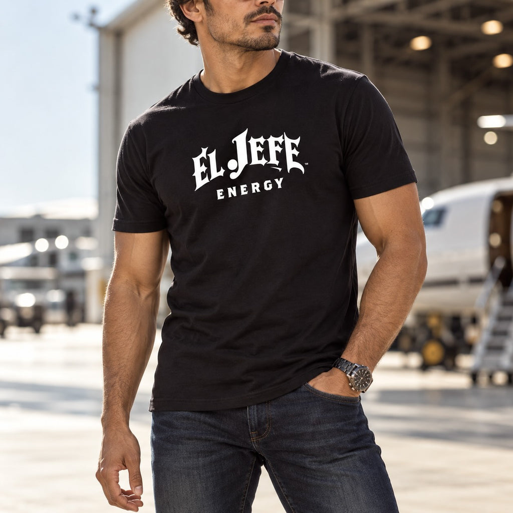 A man wearing a black crewneck t-shirt with 'EL JEFE ENERGY' printed in white gothic-style lettering, standing in front of an airplane hangar.