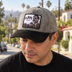 A man wearing a gray and black cap with a rectangular embroidered patch that reads 'EL JEFE ENERGY' and shows a black-and-white graphic. The background is an outdoor street scene with palm trees and buildings.