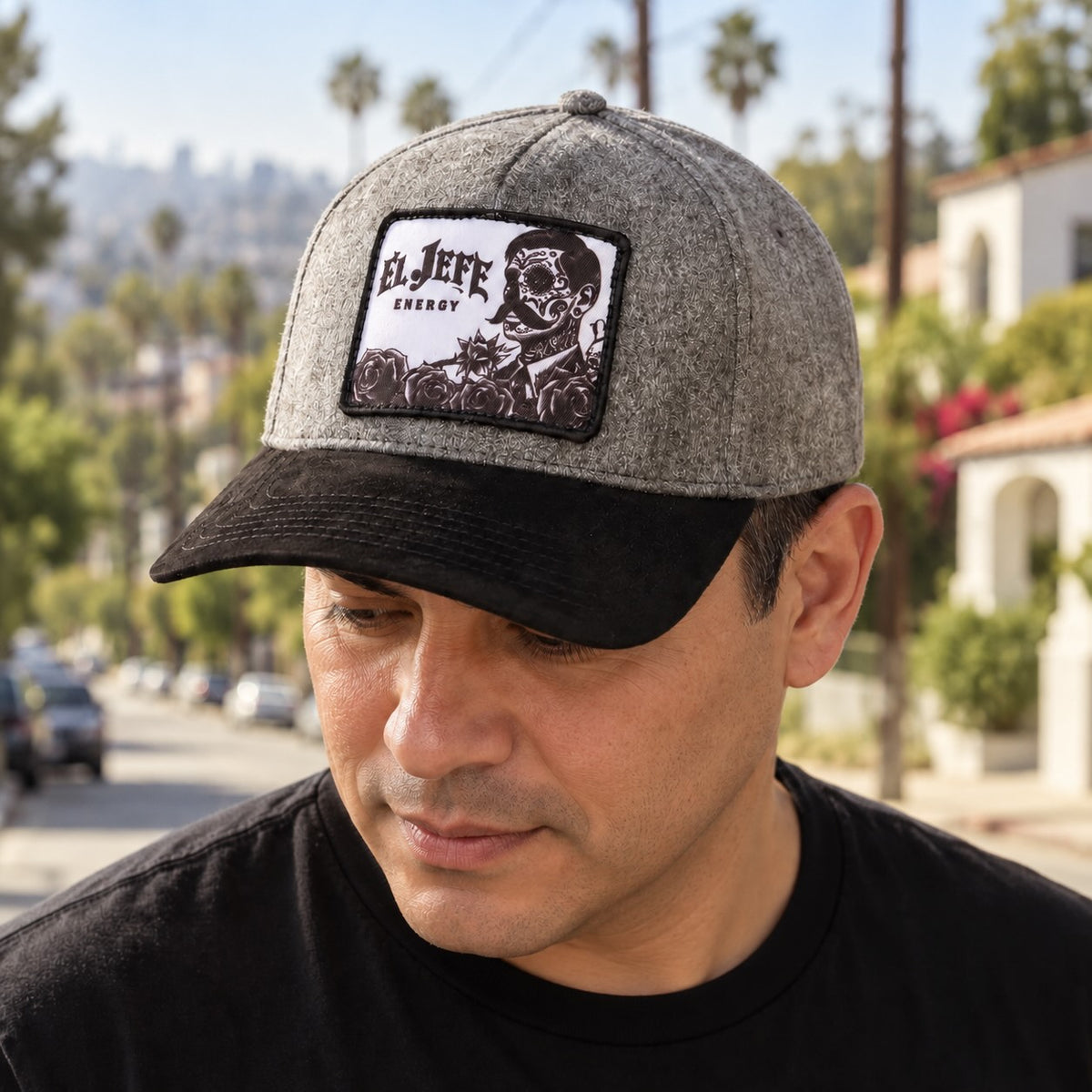 A man wearing a gray and black cap with a rectangular embroidered patch that reads 'EL JEFE ENERGY' and shows a black-and-white graphic. The background is an outdoor street scene with palm trees and buildings.