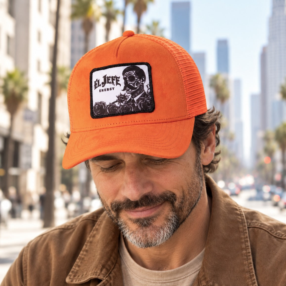 A man wearing an orange suede trucker hat with a black and white 'El Jefe Energy' patch on the front, photographed outdoors on a city street with blurred buildings and palm trees in the background.