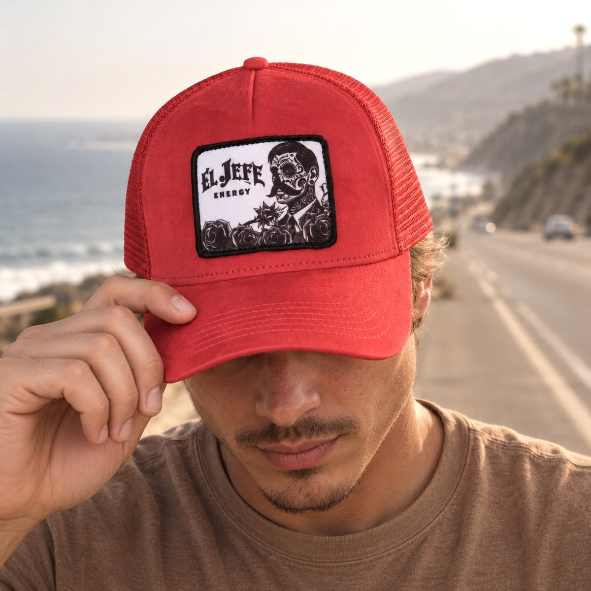 A man wearing a red suede trucker hat with a black and white 'El Jefe Energy' patch, standing on a coastal road with the ocean in the background.