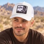 A man wearing a white suede trucker hat with a black graphic patch that reads 'EL JEFE ENERGY' and a skull illustration, photographed outdoors against a blurred desert landscape.